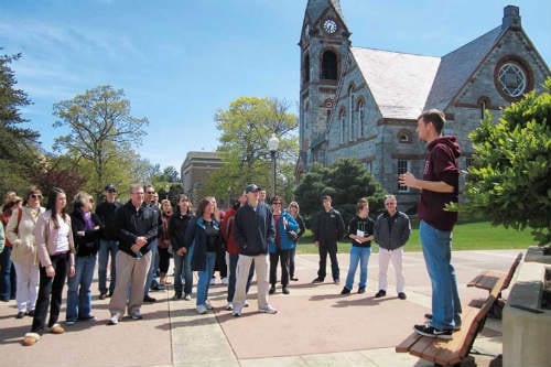 A group of students face a tour guide with a college building in the background.
