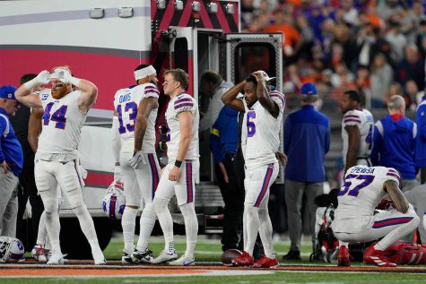 Buffalo Bills players standing in the foreground with their hands on their heads looking shocked and scared.  An ambulance behind the players preparing to take Damar Hamlin to the hospital.