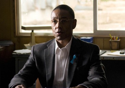 Gus Fring, an older, nicely dressed man, sitting at a desk and wearing a blue ribbon on his suit